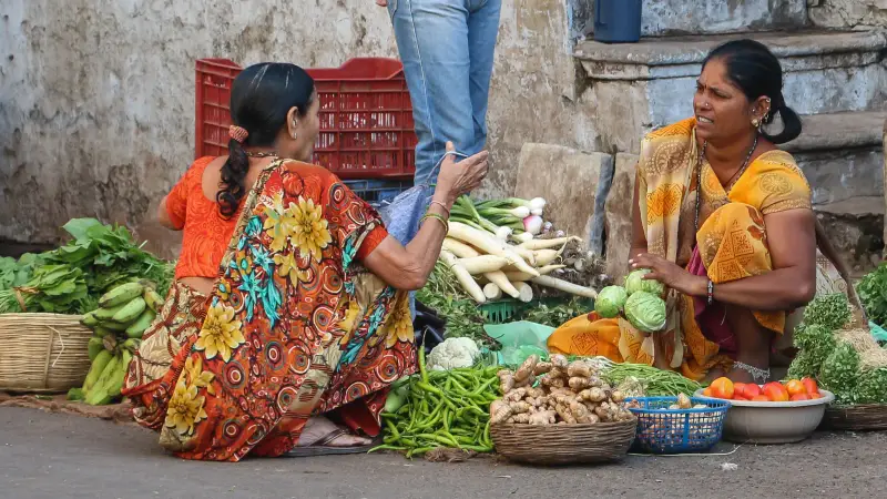 सड़क किनारे सब्ज़ी की दुकान पर दो महिलाएँ: एक पत्तागोभी उठाए सब्ज़ी बेच रही है, दूसरी ख़रीदने बैठी है; सामने टोकरियों में हरी मिर्च, अदरक, टमाटर, मूली और केले।