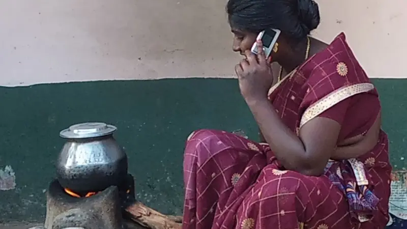 चूल्हे पर रखे बर्तन के पास बैठी साड़ी पहने एक महिला, कान से छोटा मोबाइल फ़ोन लगाकर बात करती हुई
