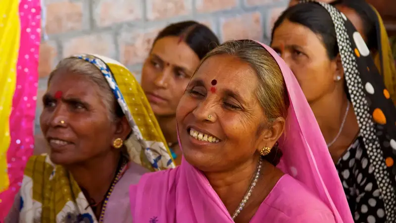 ईंट की दीवार के आगे कई ग्रामीण महिलाएँ; सामने गुलाबी साड़ी में सिर ढके एक महिला मुस्करा रही है, उसके पास पीली-सफ़ेद साड़ी में एक बुज़ुर्ग महिला, पीछे और महिलाएँ।