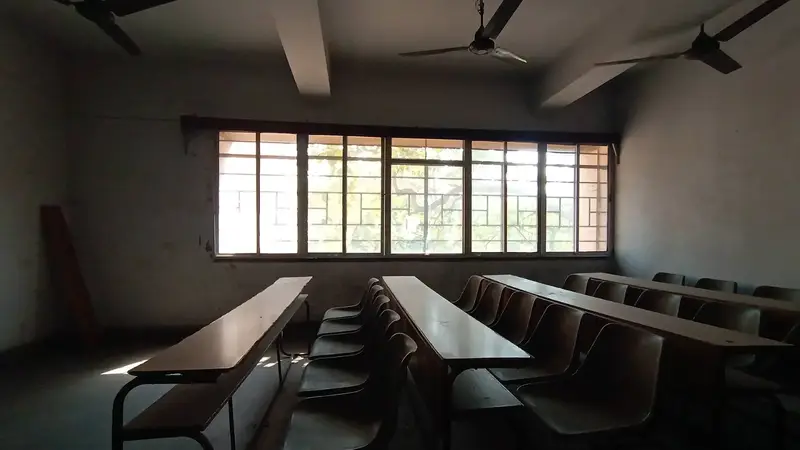 एक ख़ाली क्लासरूम: लंबी लकड़ी की मेज़ों के साथ कुर्सियों की कतारें, छत पर तीन पंखे, और पीछे की चौड़ी खिड़की से आती धूप